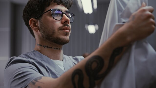 Close Up of Young Male Doctor in Glasses, Removes Protective Mask, Rubs Eyes From Fatigue, and Takes Off White Medical Coat in Hospital Locker Room. Concept of Exhaustion and Hard Work in Healthcare.