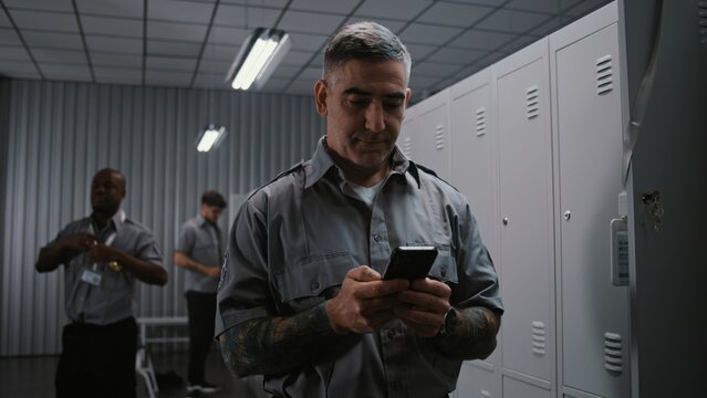 Security Officer With Arm Tattoos Checks Notifications and Social Media on Smartphone by Locker, While Other Staff Members Undress After Airport Shift. Workplace Communication and Relaxation Concept.