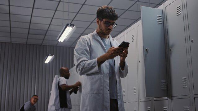 Young Male Doctor in White Coat Takes Smartphone From Metal Locker and Checks Messages, While Colleagues Undress After Medical Shift in Clinic Locker Room. Concept of Healthcare Communication.