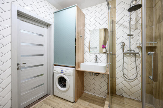 compact modern bathroom with herringbone-tiled walls, a built-in wooden cabinet, sink on a floating shelf, glass-enclosed shower, washing machine, frosted glass door, and a small flower vase for decor