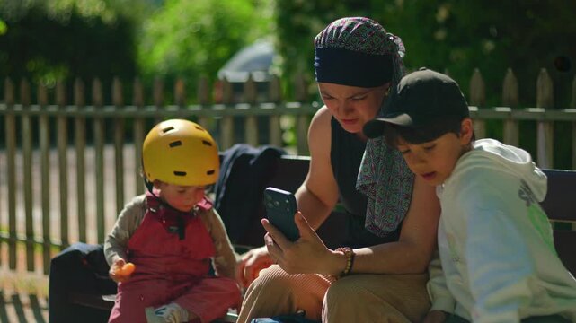 Mother undergoing chemotherapy sitting on park bench with two children, holding smartphone while handing tangerine to older son as toddler eats, candid family moment in sunlight