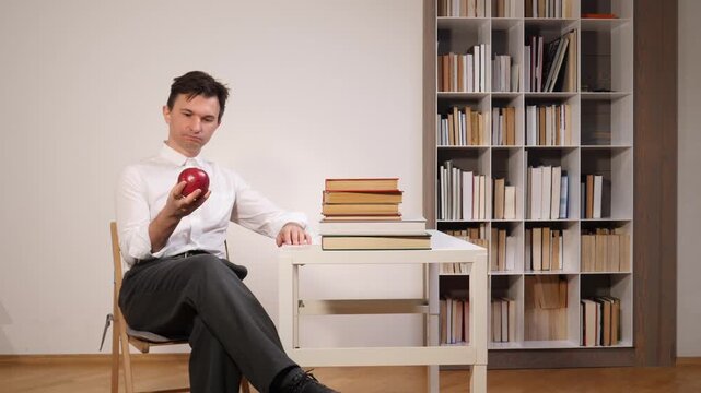 Man in a white shirt and grey trousers sitting by a desk with books, deeply thinking while holding a red apple, surrounded by a bookshelf filled with literature