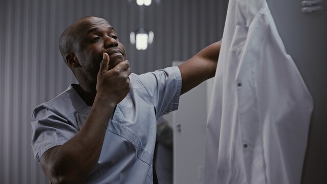 Close Up of Stressed Male African American Physician Rubbing Face in Exhaustion, With Medical Scrubs and White Coat by Grey Lockers in Clinic Locker Room Background. Stress and Burnout in Healthcare.