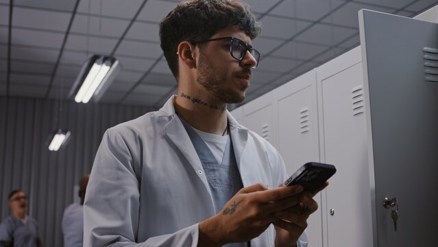 Concentrated Young Doctor With Tattoos on Arm Uses Smartphone to Study Critical Medical Reports and Lab Findings in Hospital Dressing Room After Work. Concept of Post Shift Clinical Analysis.