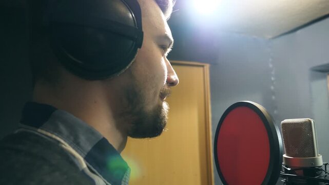 Profile of male singer in headphones singing song into the microphone at sound studio. Young man recording new melody or song. Working of creative musician. Show business concept. Slow motion