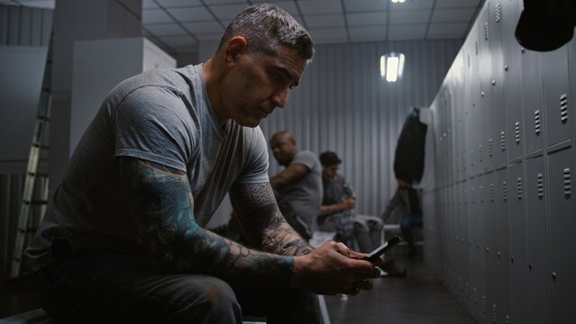 Tired Male Factory Worker Sits on Bench, Removes Orange Safety Helmet, and Checks Messages or Social Media on Smartphone in Modern Industrial Locker Room. End of Shift Routine and Digital Connection.