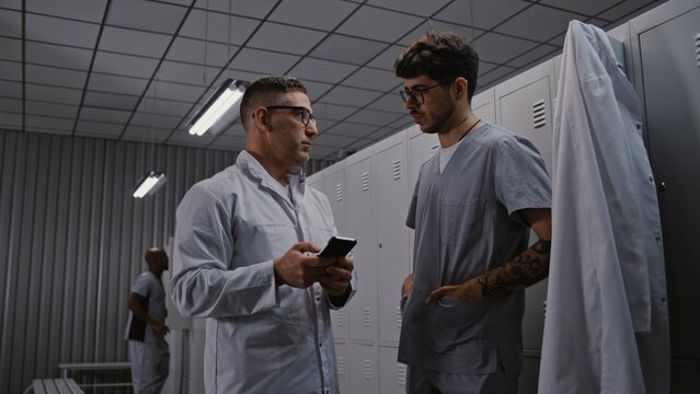 Healthcare Collaboration Concept. Senior Doctor and Younger Physician Consult Medical App on Smartphone, Intensely Discussing Treatment Options, Protocols For Severe Patient in Hospital Changing Room.