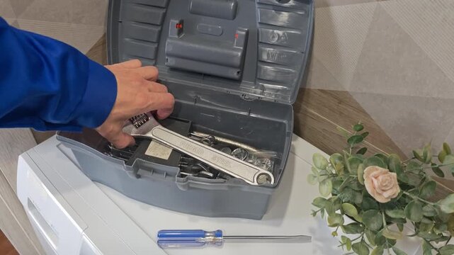 Technician repairs washing machine using tools from a gray toolbox, showcasing a wrench and screwdriver while adjusting the control panel in a domestic setting