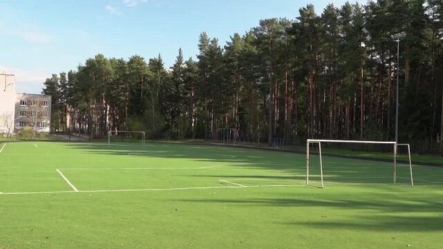 Empty soccer field at school stadium with green artificial turf, surrounded by trees and buildings, showcasing a clear blue sky and bright sunlight illuminating the scene