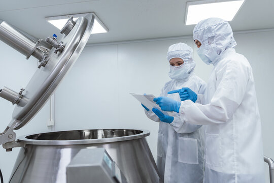 Two technicians wearing white protective suits and face masks. Professional scientists in cleanroom suits inspecting pharmaceutical manufacturing processes and operating industrial mixing equipment.