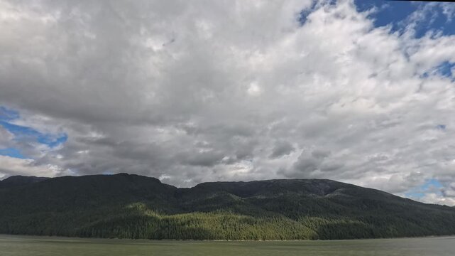 Alaska, USA - Mountain range covered in dense forest along the water in Misty Fjords. Cloudy sky with some blue patches above the landscape.