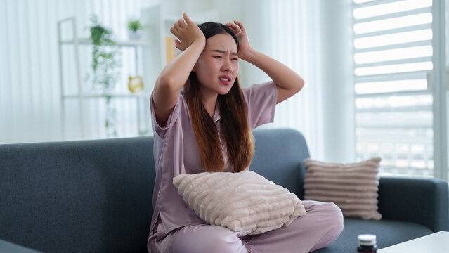young Asian woman suffering headache migraine pain sitting on sofa holding head stressed frustrated feeling unwell depressed at home living room mental health