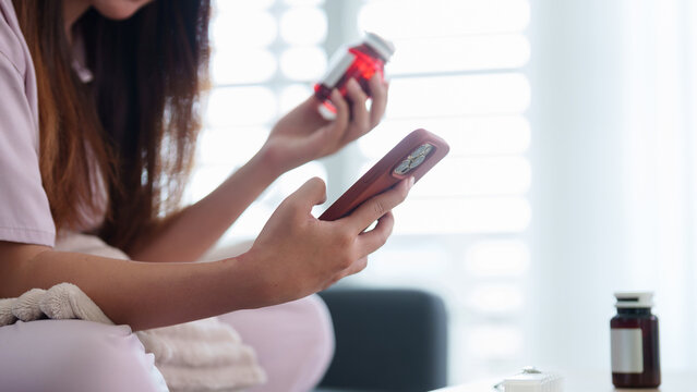 Asian women using smartphone reading medicine bottle label checking prescription information sitting home with vitamin supplement pill healthcare wellness