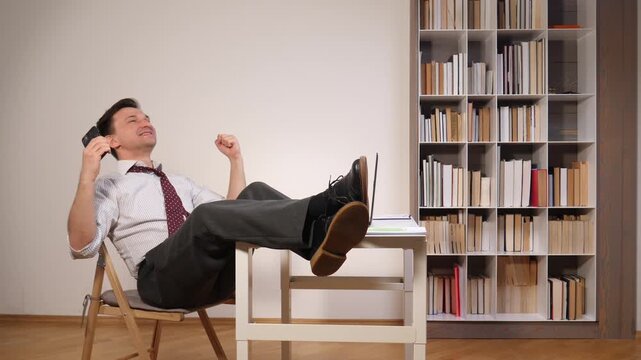 Content businessman relaxing in office chair with feet up on a messy desk next to a laptop and bookshelf, celebrating an achievement or successful project