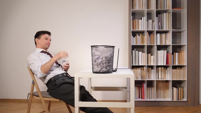 Businessman sitting at a desk and playing with crumpled paper, throwing it into a waste bin, highlighting themes of boredom, wasting time, and inefficiency at work