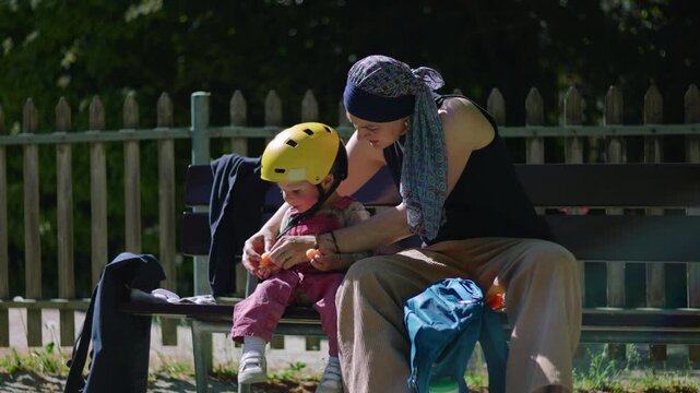 Mother undergoing chemotherapy sitting on park bench feeding toddler, headscarf parent sharing snack in sunlight, tender caregiving moment showing connection and resilience