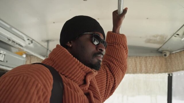 Black man in orange knit sweater holding handrail inside bus. Quiet portrait captures commuter as designer checking route, student lost in thought, musician rehearsing lyrics silently, photographer