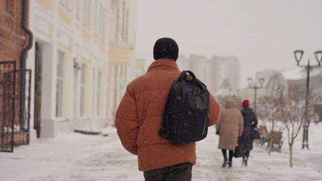 Man walking away snowy street, downtown brick facades, puffy coat and shoulder bag, footsteps on icy sidewalk, soft falling snow, muted palette, solitary commute and contemplative mood