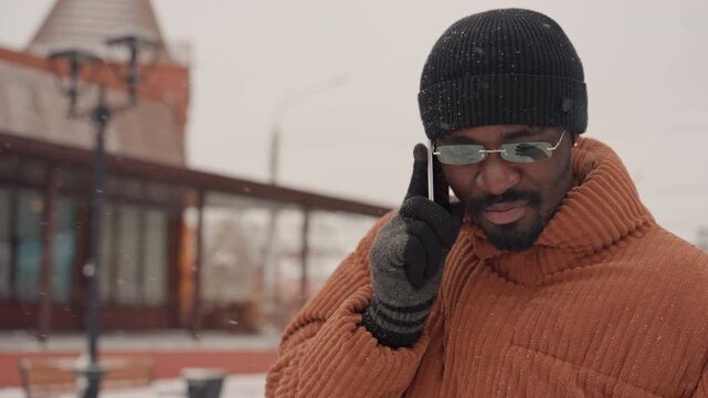 Black man talking on phone in brown knit coat and black beanie, wearing glasses and wool gloves, backpack over shoulder, walking through snowy city street with falling snow, expressive smile, candid