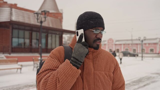 Black man in orange coat talking on phone at snowy plaza, wearing black beanie and wool gloves, backpack over shoulder, pensive expression, soft snowfall, empty benches and lamppost, muted brick