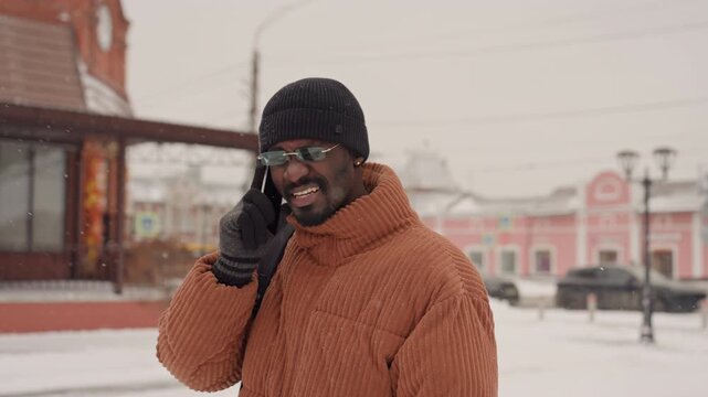 Black man orange coat on phone in snowy plaza beanie and glasses, gesturing while walking, pausing to look upward, checking directions, making business call, visible backpack, breath in cold air,