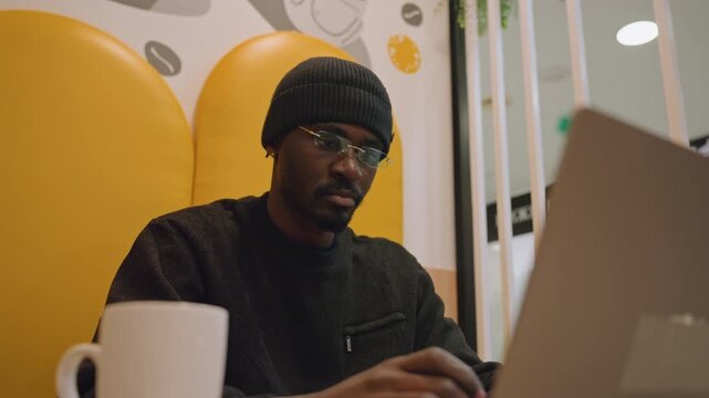 Black student studying laptop in cafe, beanie and glasses, notepad and pen, research notes spread on table, warm mall lighting, quiet concentration, exam prep mood, slow thoughtful typing