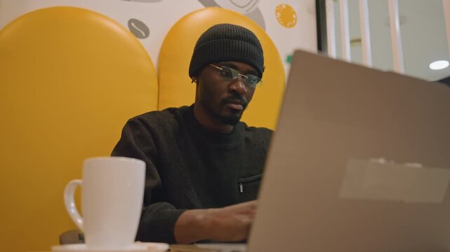 Black entrepreneur planning startup on laptop, beanie and glasses, coffee mug at foreground, yellow booth backdrop, sketching notes and pen, focused brainstorming for pitch, investor prep vibe, modern