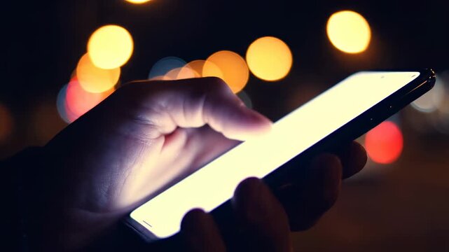 Close-up of a hand using a smartphone at night with a bright white screen and city bokeh background for mobile app marketing and digital communication concepts.