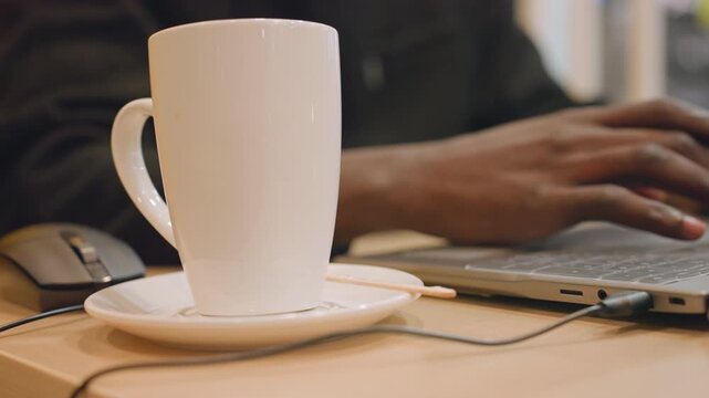 White mug foreground black hands typing near laptop and mouse on wooden desk, soft sunlight through window, writer drafting notes with notebook and pen, ceramic saucer and spoon visible, quiet