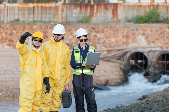 Workers in yellow suits and hard hats kneel by a river to collect water samples for testing at industrial site