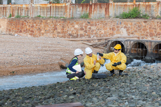 Workers in yellow suits and hard hats kneel by a river to collect water samples for testing at industrial site