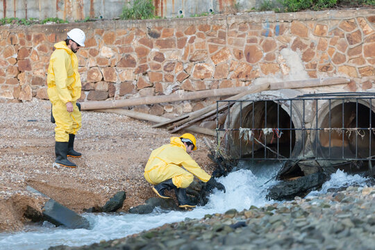 Workers in yellow suits and hard hats kneel by a river to collect water samples for testing at industrial site