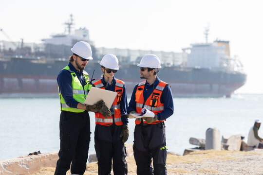 Workers discuss plans at a port with a cargo ship in the background