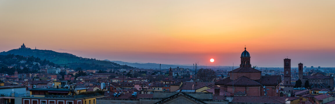 Panoramic sunset view over the skyline of Bologna, Italy, with the Santuario della Madonna di San Luca visible on the hill.