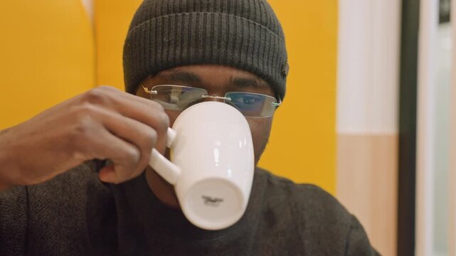 Black man sipping coffee at table during study break, wearing beanie and glasses, yellow booth setting, reading notes and checking laptop, pensive expression, quiet mall cafe atmosphere, soft bokeh