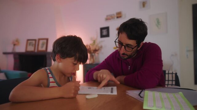 Father helping son with homework at table in evening, supportive parenting and home education routine, guidance and concentration in calm family learning moment