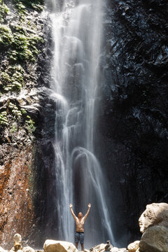 Man tourist with raised arms under powerful jungle waterfall, feeling energy in tropical nature