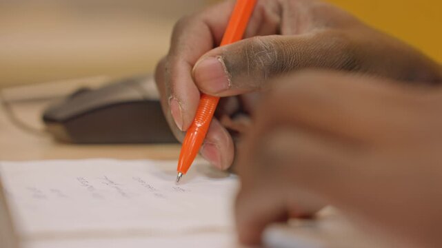 Closeup hands writing orange pen notes on desk concentrated student capturing key points beside computer mouse, warm desk lighting, textured stationery and organized paper, calm exam preparation