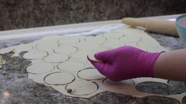 Close up of hands in pink gloves cutting round dough circles for homemade dumplings pelmeni on floured table with rolling pin and bowl