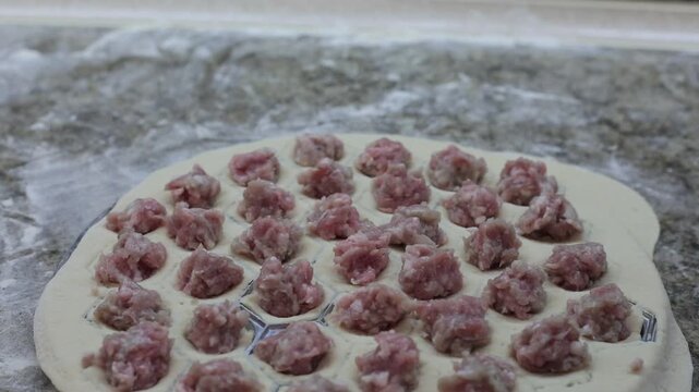  woman placing small portions of raw minced meat filling onto rows of dough circles on a floured countertop while making traditional homemade pelmeni or dumplings by hand