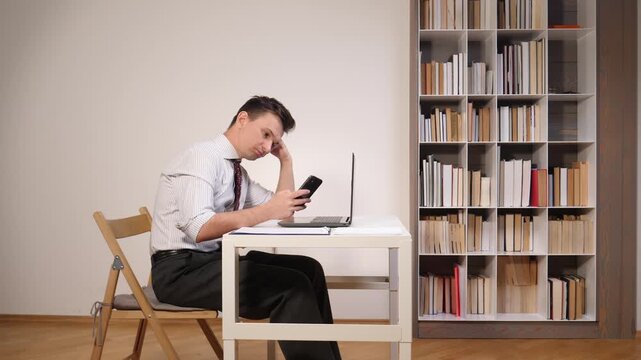 Man in a formal shirt feeling bored and procrastinating by checking his smartphone instead of working on a laptop at a small desk in a room with a bookshelf