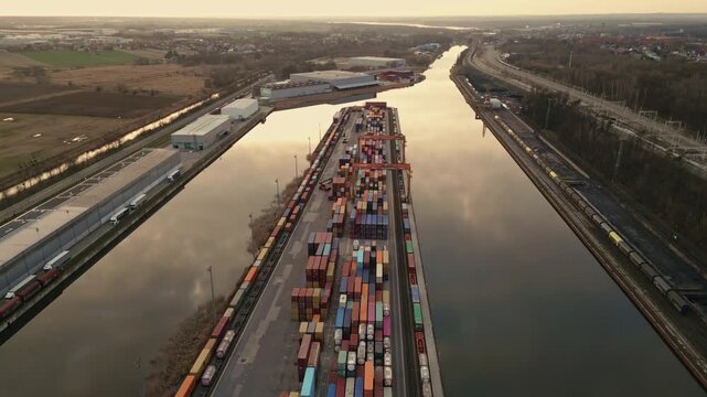 Aerial orbital drone flight circling the Gliwice inland port container terminal during sunset. View of colorful containers, cranes, and Gliwice Canal with reflections in the water.