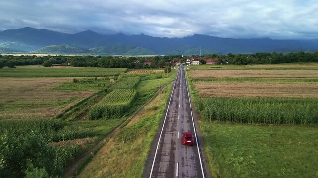 Aerial forward and rising drone flight overtaking a red car on a road towards Cartisoara village and the Fagaras Mountains in Romania. Scenic rural landscape with fields and mountain range.