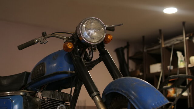 Blue classic motorcycle with a round headlight parked indoors, showing its weathered paint, handlebars, and front fender, representing nostalgia, restoration, and old fashioned travel