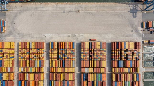 Aerial view of a shipping container yard with colorful containers neatly arranged in rows, adjacent to a dock with water on one side.