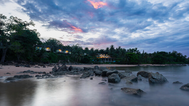A serene tropical beach scene at sunset featuring smooth long-exposure water flowing around dark rocks in the foreground. Lush palm trees and hillside villas glow warmly in the background under a dram