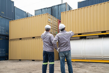 Fototapeta premium Two engineers workers in hard hats inspect stacked shipping containers at a freight yard, discussing logistics and safety procedures.