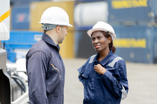 Two engineers industrial workers in helmets and coveralls talk at a shipping yard with stacked cargo containers in the background.