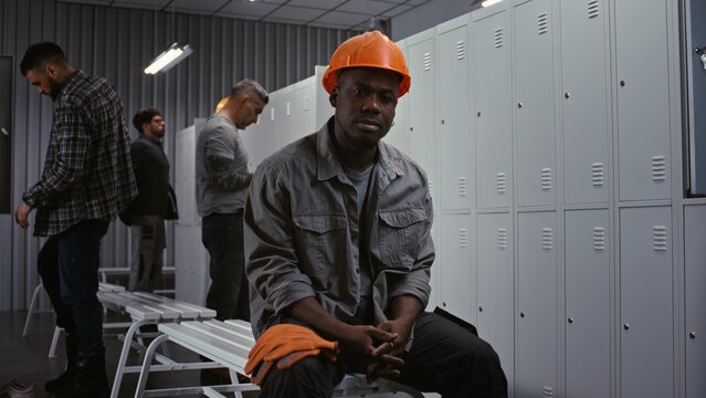 African American Factory Worker Sits on Bench, Wearing Orange Safety Helmet and Grey Jacket, With Metal Lockers and Diverse Colleagues in Background. Industrial Camaraderie and Diversity. Portrait.
