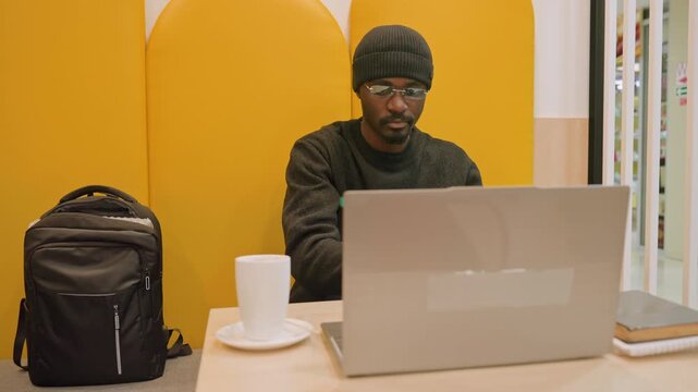 Man packing laptop into backpack, yellow booth cafe scene, steaming mug and notebook nearby, beanie and headphones on, zipping bag and securing laptop for quick exit, urban commute vibe and focused